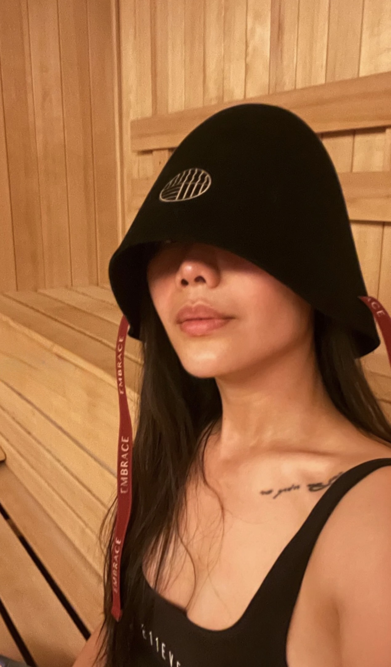 A close portrait of a woman wearing the black Embrace sauna hat with its EMBRACE straps