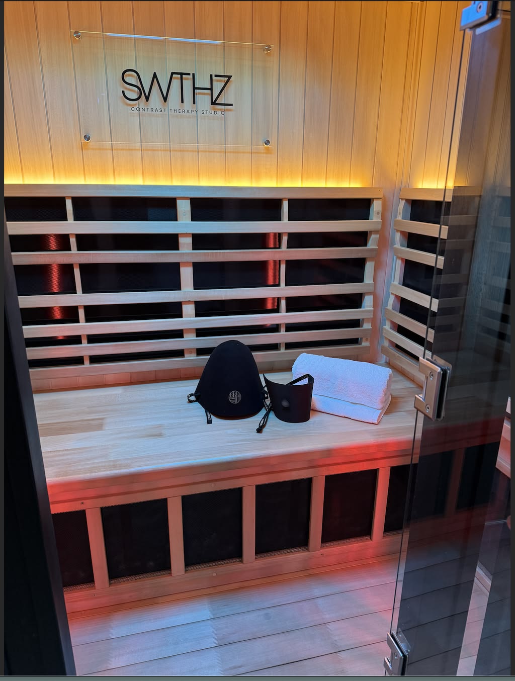 The Embrace sauna hat resting on a cedar bench beside rolled towels in a warmly lit studio sauna