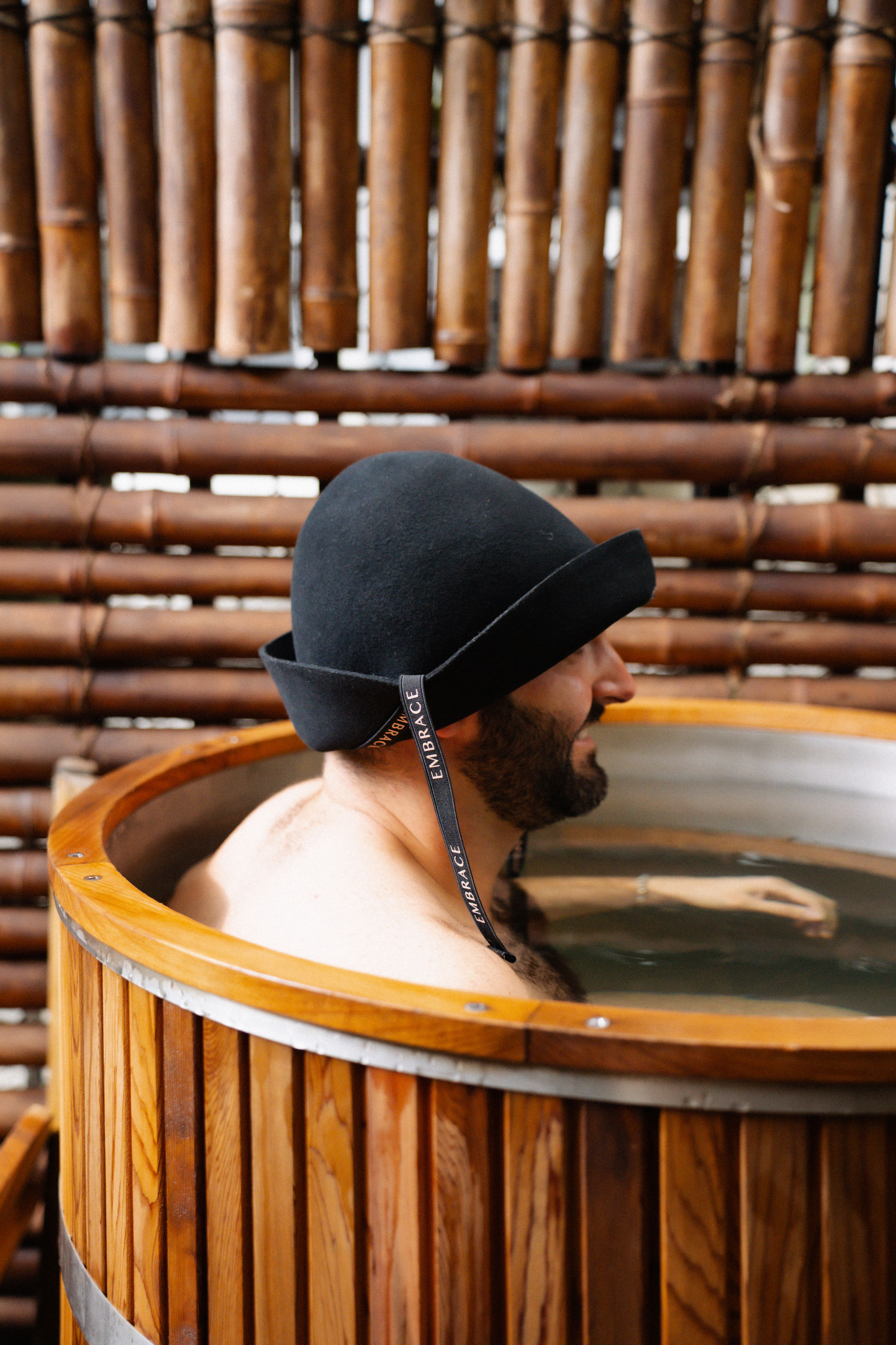 A man cooling down in a wooden cold plunge, wearing the black Embrace sauna hat in profile