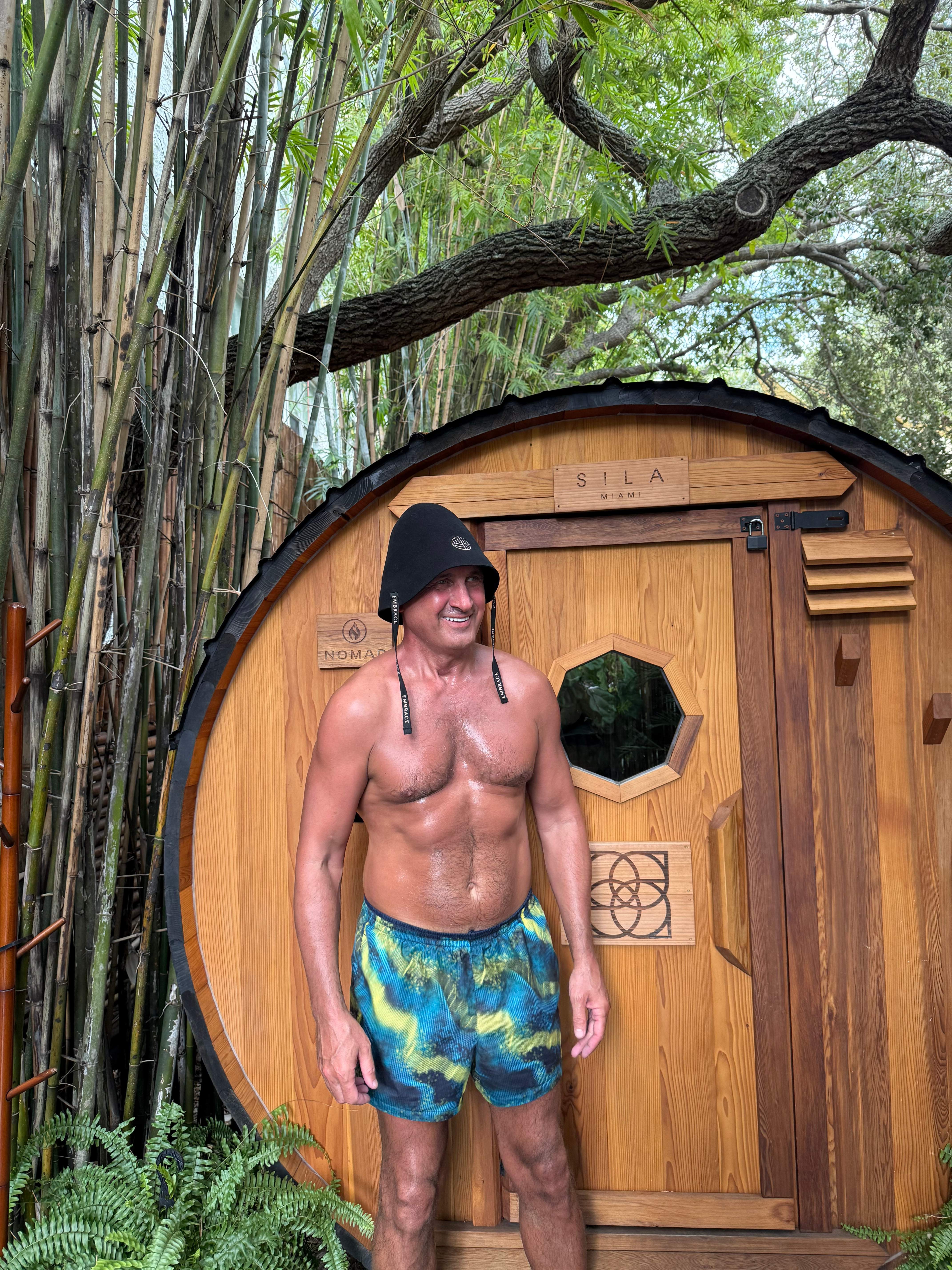 A smiling man outside a barrel sauna surrounded by bamboo, wearing the Embrace sauna hat