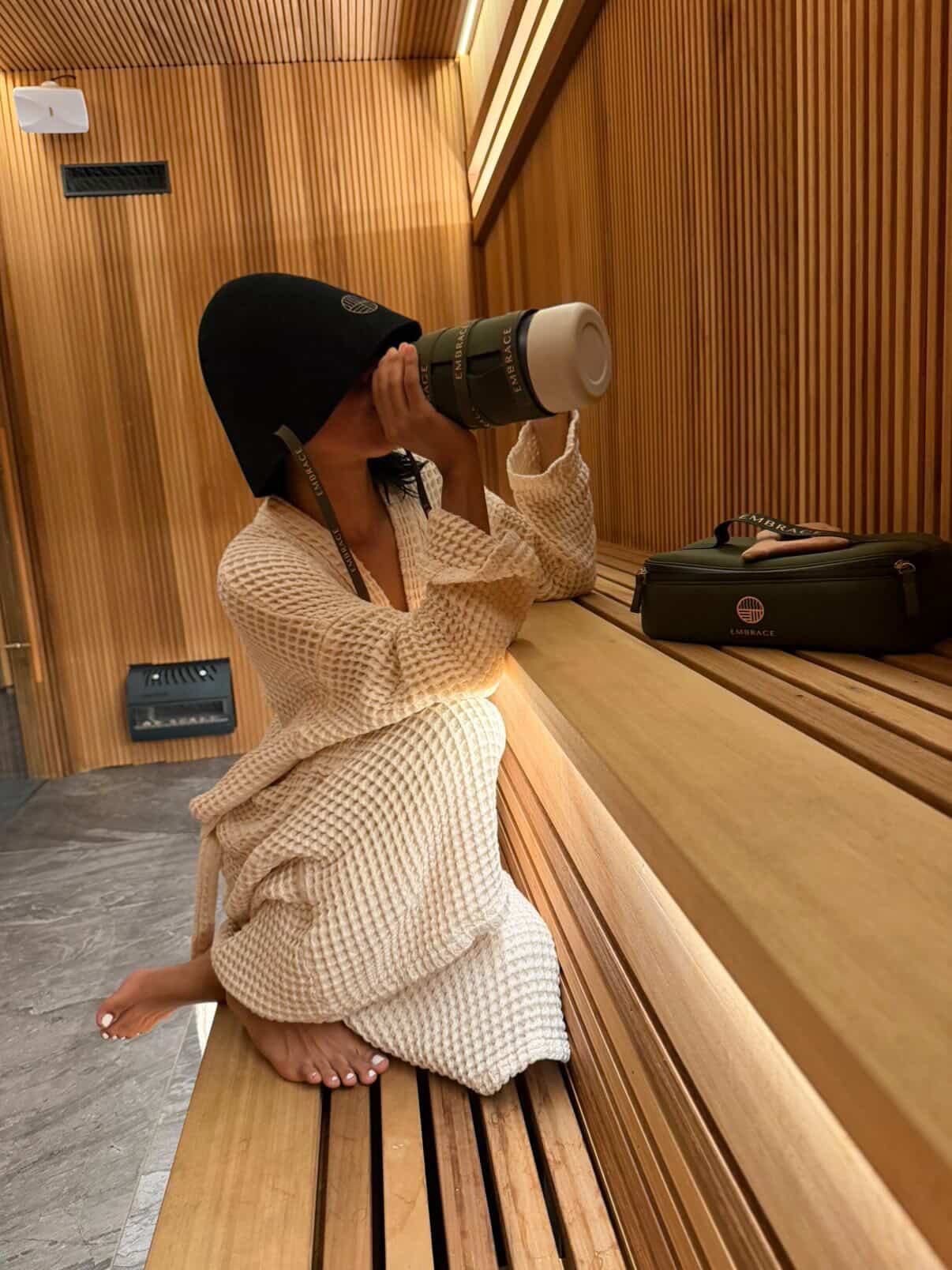 A woman in a waffle robe kneeling on a sauna bench, sipping from an Embrace bottle with the sauna hat on